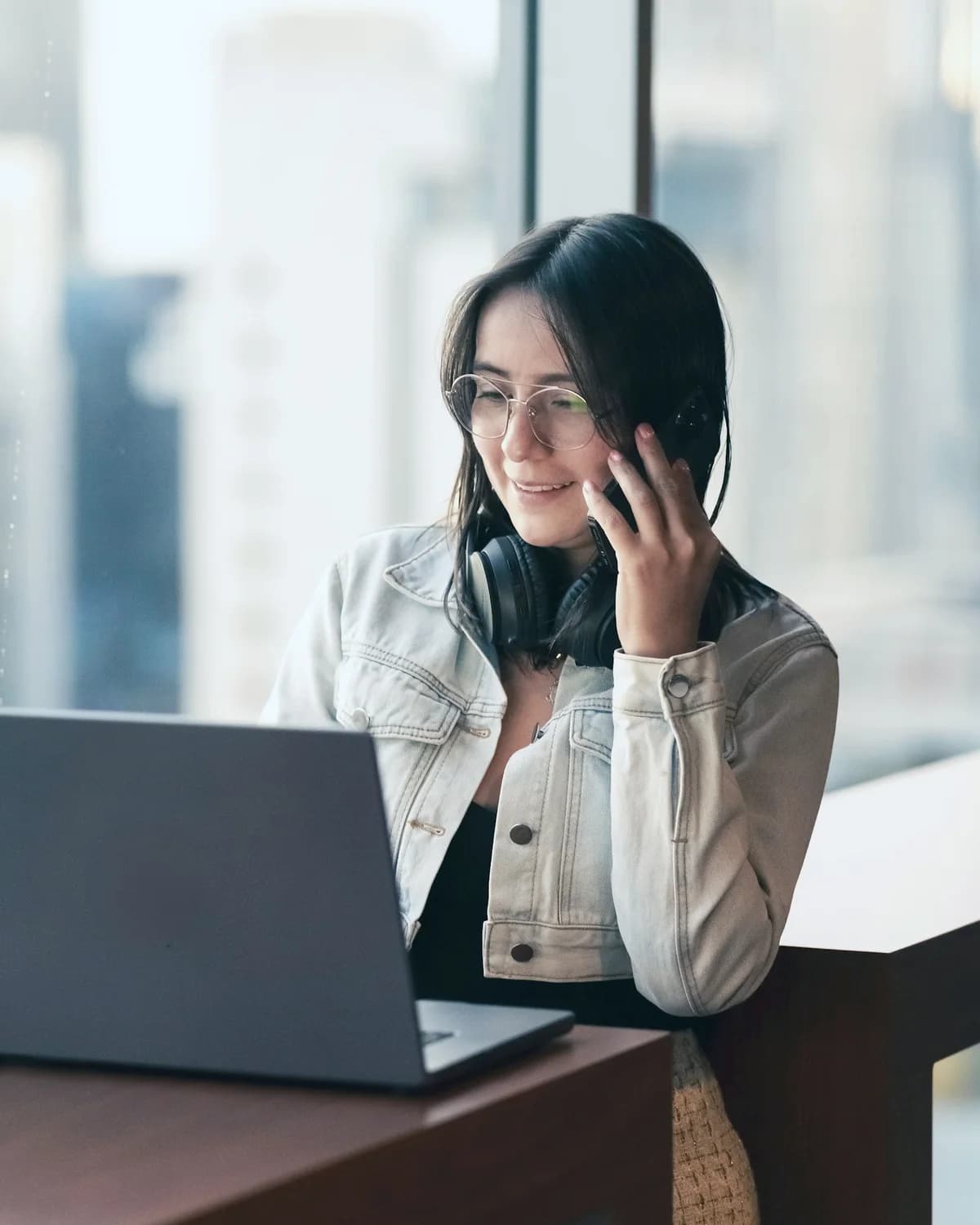 Mulher sorrindo ao celular, trabalhando no notebook perto da janela